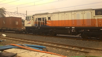 A freight train with a locomotive painted in white, orange, and blue, pulling a cargo container. The train is on a set of railway tracks with electric lines overhead and some industrial elements in the background. A few cables and parts of a motorcycle are visible in the foreground.