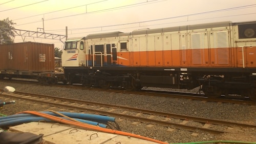 A freight train with a locomotive painted in white, orange, and blue, pulling a cargo container. The train is on a set of railway tracks with electric lines overhead and some industrial elements in the background. A few cables and parts of a motorcycle are visible in the foreground.