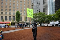 A person wearing a black hat and jacket holds a bright green sign that reads 'Say No to GMO' in a city setting. In the background, a group of people is gathered near an old-style city bus. Large trees and the facade of a multi-story building add to the urban atmosphere.