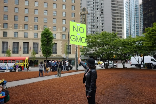 A person wearing a black hat and jacket holds a bright green sign that reads 'Say No to GMO' in a city setting. In the background, a group of people is gathered near an old-style city bus. Large trees and the facade of a multi-story building add to the urban atmosphere.