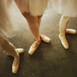 Three ballet dancers' legs are shown wearing pointe shoes on a dark floor. The dancers are dressed in light-colored tutus, and their shoes have a soft pink hue. The composition focuses on the elegance and poise of the dancers' feet.