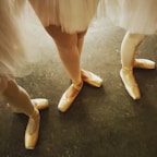 Three ballet dancers' legs are shown wearing pointe shoes on a dark floor. The dancers are dressed in light-colored tutus, and their shoes have a soft pink hue. The composition focuses on the elegance and poise of the dancers' feet.