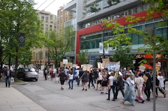 A group of people are marching on an urban street, holding up signs and banners. The area is lined with trees and various shops, with a modern building in the background. Vehicles are visible parked along the street and there are other pedestrians walking nearby.