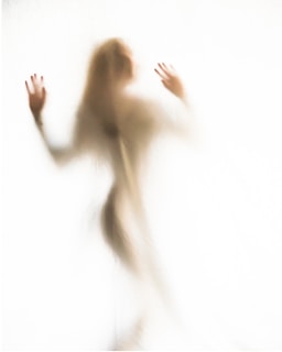 A shadowy, blurred figure of a person is seen through an opaque surface with their hands slightly raised. The image has a soft and ethereal quality, with indistinct features and a ghostly appearance.