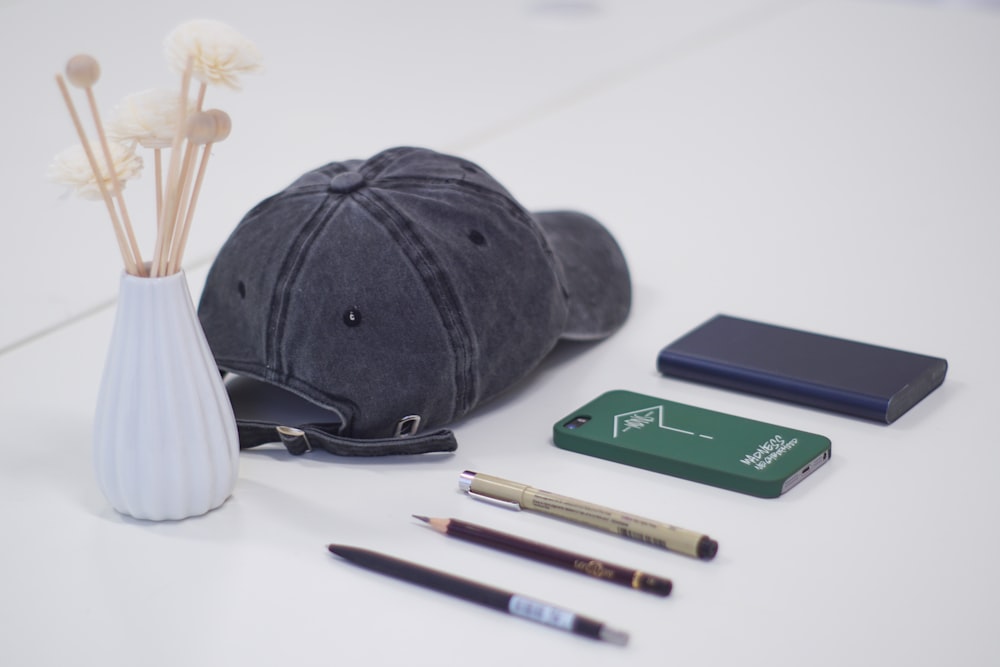 gray fitted cap, three pen, smartphone, and blue power bank on white surface