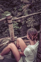 A person is sitting on a wooden platform surrounded by a natural environment. The person is wearing casual clothing and has their hair tied back, looking towards the lush greenery in the background. The setting includes wooden railings and dense foliage, indicating a serene, outdoor location.
