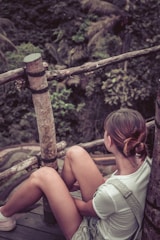 A person is sitting on a wooden platform surrounded by a natural environment. The person is wearing casual clothing and has their hair tied back, looking towards the lush greenery in the background. The setting includes wooden railings and dense foliage, indicating a serene, outdoor location.