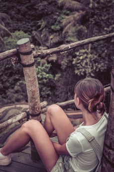 A person is sitting on a wooden platform surrounded by a natural environment. The person is wearing casual clothing and has their hair tied back, looking towards the lush greenery in the background. The setting includes wooden railings and dense foliage, indicating a serene, outdoor location.