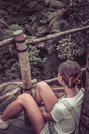 A person is sitting on a wooden platform surrounded by a natural environment. The person is wearing casual clothing and has their hair tied back, looking towards the lush greenery in the background. The setting includes wooden railings and dense foliage, indicating a serene, outdoor location.