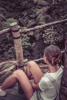 A person is sitting on a wooden platform surrounded by a natural environment. The person is wearing casual clothing and has their hair tied back, looking towards the lush greenery in the background. The setting includes wooden railings and dense foliage, indicating a serene, outdoor location.