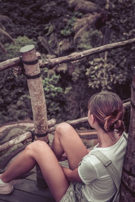 A person is sitting on a wooden platform surrounded by a natural environment. The person is wearing casual clothing and has their hair tied back, looking towards the lush greenery in the background. The setting includes wooden railings and dense foliage, indicating a serene, outdoor location.