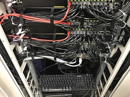 A network server rack filled with neatly organized cables. There are numerous black and red ethernet cables bundled together using cable ties. A keyboard and computer are placed at the bottom of the rack, with various networking equipment above them, showing multiple ports and blinking green lights indicating active connections.