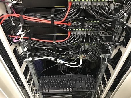 A network server rack filled with neatly organized cables. There are numerous black and red ethernet cables bundled together using cable ties. A keyboard and computer are placed at the bottom of the rack, with various networking equipment above them, showing multiple ports and blinking green lights indicating active connections.