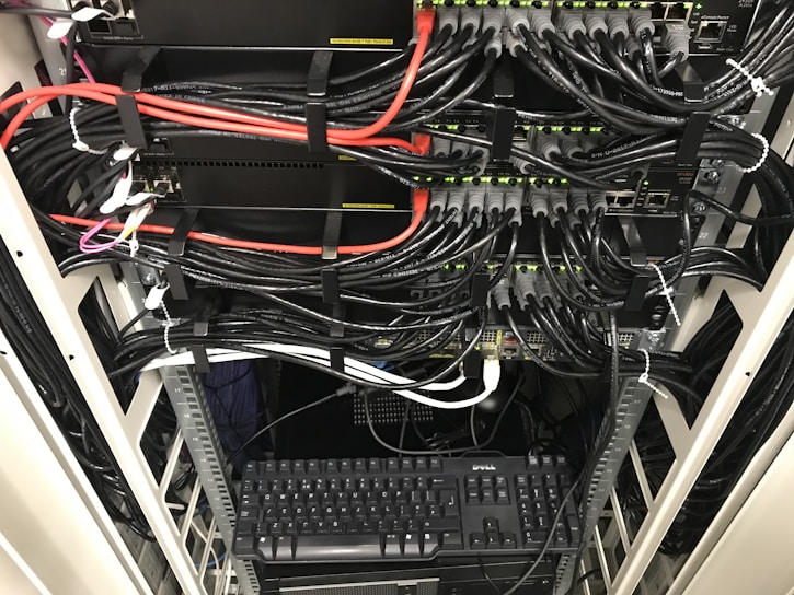 A network server rack filled with neatly organized cables. There are numerous black and red ethernet cables bundled together using cable ties. A keyboard and computer are placed at the bottom of the rack, with various networking equipment above them, showing multiple ports and blinking green lights indicating active connections.