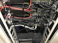 A network server rack filled with neatly organized cables. There are numerous black and red ethernet cables bundled together using cable ties. A keyboard and computer are placed at the bottom of the rack, with various networking equipment above them, showing multiple ports and blinking green lights indicating active connections.