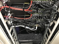 A network server rack filled with neatly organized cables. There are numerous black and red ethernet cables bundled together using cable ties. A keyboard and computer are placed at the bottom of the rack, with various networking equipment above them, showing multiple ports and blinking green lights indicating active connections.