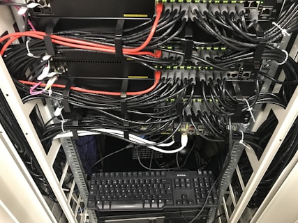 A network server rack filled with neatly organized cables. There are numerous black and red ethernet cables bundled together using cable ties. A keyboard and computer are placed at the bottom of the rack, with various networking equipment above them, showing multiple ports and blinking green lights indicating active connections.