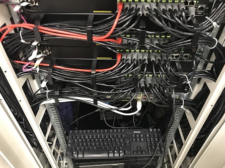 A network server rack filled with neatly organized cables. There are numerous black and red ethernet cables bundled together using cable ties. A keyboard and computer are placed at the bottom of the rack, with various networking equipment above them, showing multiple ports and blinking green lights indicating active connections.