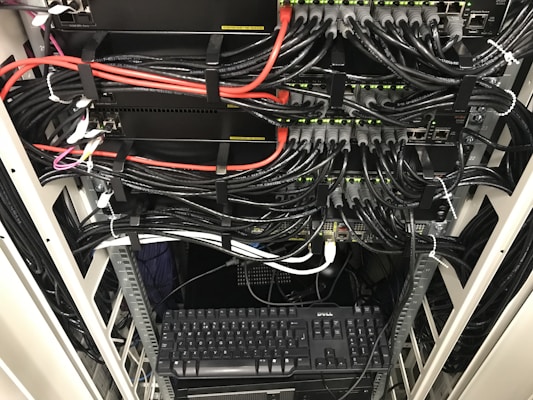 A network server rack filled with neatly organized cables. There are numerous black and red ethernet cables bundled together using cable ties. A keyboard and computer are placed at the bottom of the rack, with various networking equipment above them, showing multiple ports and blinking green lights indicating active connections.