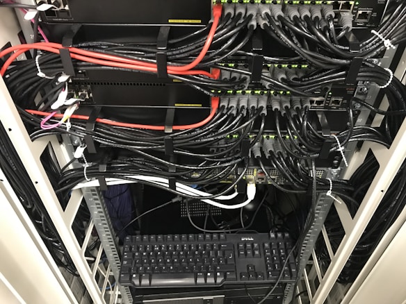 A network server rack filled with neatly organized cables. There are numerous black and red ethernet cables bundled together using cable ties. A keyboard and computer are placed at the bottom of the rack, with various networking equipment above them, showing multiple ports and blinking green lights indicating active connections.