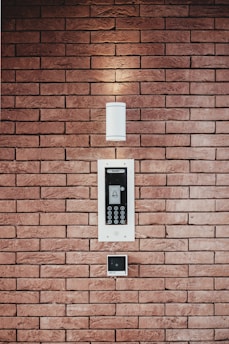 A brick wall serves as the background for an electronic intercom system, which features a sleek black and silver panel with a digital display and numeric keypad. Above the intercom, a modern light fixture emits a soft glow, creating a well-lit area. Below the intercom, a small square device is mounted on the wall.