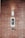 A brick wall serves as the background for an electronic intercom system, which features a sleek black and silver panel with a digital display and numeric keypad. Above the intercom, a modern light fixture emits a soft glow, creating a well-lit area. Below the intercom, a small square device is mounted on the wall.