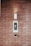 A brick wall serves as the background for an electronic intercom system, which features a sleek black and silver panel with a digital display and numeric keypad. Above the intercom, a modern light fixture emits a soft glow, creating a well-lit area. Below the intercom, a small square device is mounted on the wall.