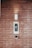 A brick wall serves as the background for an electronic intercom system, which features a sleek black and silver panel with a digital display and numeric keypad. Above the intercom, a modern light fixture emits a soft glow, creating a well-lit area. Below the intercom, a small square device is mounted on the wall.