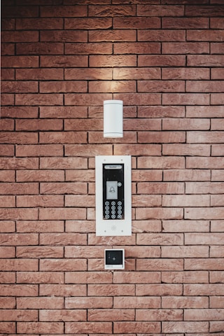 A brick wall serves as the background for an electronic intercom system, which features a sleek black and silver panel with a digital display and numeric keypad. Above the intercom, a modern light fixture emits a soft glow, creating a well-lit area. Below the intercom, a small square device is mounted on the wall.