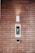 A brick wall serves as the background for an electronic intercom system, which features a sleek black and silver panel with a digital display and numeric keypad. Above the intercom, a modern light fixture emits a soft glow, creating a well-lit area. Below the intercom, a small square device is mounted on the wall.