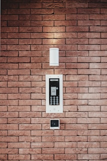 A brick wall serves as the background for an electronic intercom system, which features a sleek black and silver panel with a digital display and numeric keypad. Above the intercom, a modern light fixture emits a soft glow, creating a well-lit area. Below the intercom, a small square device is mounted on the wall.