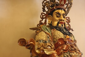 A detailed and ornate statue of a warrior, featuring intricate armor with gold and bronze colors. The figure has a fierce expression, with large eyes and a black beard. The headpiece is complex, adorned with decorative elements that include curls and floral patterns. The statue's clothing incorporates a mix of textures and materials, giving it a grand, ceremonial appearance.