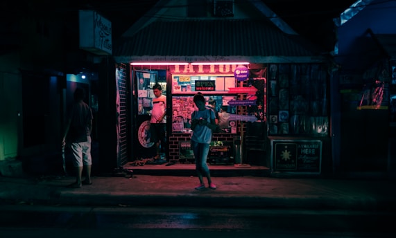 A nighttime street scene features a tattoo parlor illuminated by neon lights. The shop facade is adorned with posters and signs, and there are several people visible, one standing in the entrance and another walking along the sidewalk. The atmosphere is urban and atmospheric, with dimly lit surroundings contrasting with the vibrant glow from the shop.