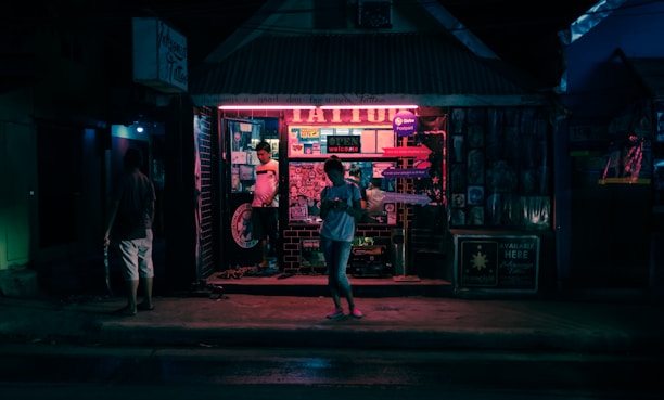 A nighttime street scene features a tattoo parlor illuminated by neon lights. The shop facade is adorned with posters and signs, and there are several people visible, one standing in the entrance and another walking along the sidewalk. The atmosphere is urban and atmospheric, with dimly lit surroundings contrasting with the vibrant glow from the shop.