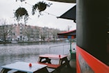 An outdoor seating area by a river features red and blue picnic tables. The scene includes a view of a waterfront with residential buildings on the opposite side. Hanging plants and string lights adorn the area, adding a cozy atmosphere.
