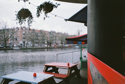 An outdoor seating area by a river features red and blue picnic tables. The scene includes a view of a waterfront with residential buildings on the opposite side. Hanging plants and string lights adorn the area, adding a cozy atmosphere.
