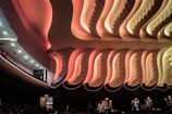 An intricately designed interior ceiling with ornate, wavy patterns and warm lighting in shades of pink and orange. The ceiling design is highlighted by soft, gradient lighting, creating an elegant and dramatic effect. Below, silhouettes of people are visible, suggesting a public space such as a theater or auditorium. Exit signs illuminate the edges, indicating multiple exits are present.