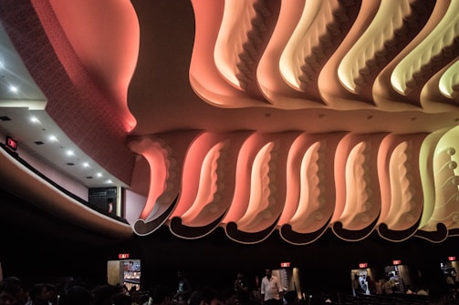 An intricately designed interior ceiling with ornate, wavy patterns and warm lighting in shades of pink and orange. The ceiling design is highlighted by soft, gradient lighting, creating an elegant and dramatic effect. Below, silhouettes of people are visible, suggesting a public space such as a theater or auditorium. Exit signs illuminate the edges, indicating multiple exits are present.