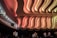 An intricately designed interior ceiling with ornate, wavy patterns and warm lighting in shades of pink and orange. The ceiling design is highlighted by soft, gradient lighting, creating an elegant and dramatic effect. Below, silhouettes of people are visible, suggesting a public space such as a theater or auditorium. Exit signs illuminate the edges, indicating multiple exits are present.