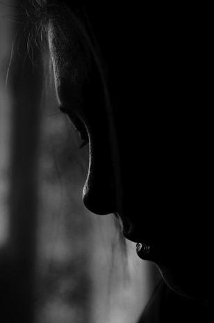 A monochromatic profile of a person shown in a close-up view with strong contrast. The lighting accentuates the contours of the face, particularly the nose and lips, creating a dramatic and mysterious atmosphere.