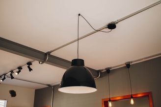 A modern ceiling light fixture with a large black lampshade is centered in the image. The ceiling has metal conduit pipes with additional smaller industrial-style light fixtures attached. The room has neutral gray walls, and mirrors with wooden frames can be seen along with two hanging light bulbs beside the central lamp.