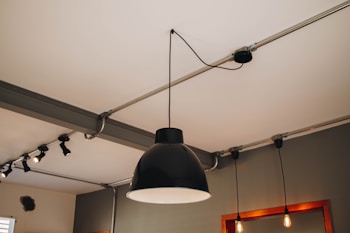 A modern ceiling light fixture with a large black lampshade is centered in the image. The ceiling has metal conduit pipes with additional smaller industrial-style light fixtures attached. The room has neutral gray walls, and mirrors with wooden frames can be seen along with two hanging light bulbs beside the central lamp.