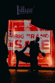 Silhouetted figures create a dramatic contrast against an illuminated store window displaying a 'Big Brand Sale' advertisement. The scene is dominated by dark shadows and vibrant red hues from the sign, highlighting the energetic and busy vibe of a city at night.