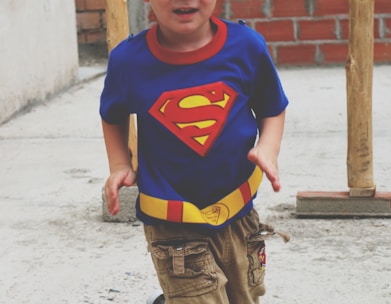 A young child is wearing a blue t-shirt with a red and yellow Superman logo and a utility belt design. The child is in motion, with arms slightly bent and appearing to be running or playing. The background shows a concrete surface and a brick wall, with wooden beams visible.