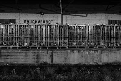 A row of metal cages lined up on a platform, possibly for storage or transport. The background features an industrial wall with small windows and a sign that reads 'RAUCHVERBOT,' indicating a no smoking zone. The overall scene appears to be at a loading dock or similar industrial setting.