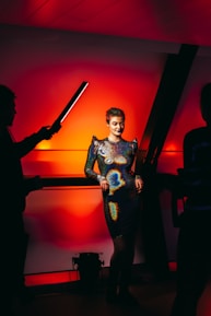 A person stands confidently in a glittering, form-fitting dress with a futuristic design, set against a vibrant, red-lit background. The scene is framed by silhouetted figures holding light equipment, indicating a photo or video shoot setting.
