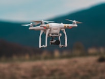 A quadcopter drone is captured in mid-air flight against a blurred mountainous backdrop. The drone is white with gold accents and is equipped with a camera, highlighting its capabilities for aerial photography.