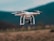 A quadcopter drone is captured in mid-air flight against a blurred mountainous backdrop. The drone is white with gold accents and is equipped with a camera, highlighting its capabilities for aerial photography.