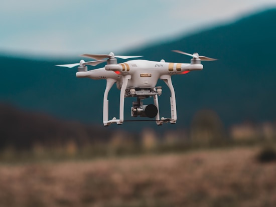 A quadcopter drone is captured in mid-air flight against a blurred mountainous backdrop. The drone is white with gold accents and is equipped with a camera, highlighting its capabilities for aerial photography.