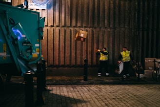 Two workers in high-visibility jackets are handling packages near a large garbage truck. One person is throwing a cardboard box towards the truck while the other stands holding bags. The ground is cobblestone, and the setting appears to be a dimly lit urban street at night.